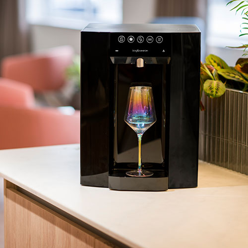 Active B&O E6 table top water cooler in an office environment with wine glass in dispensing area.